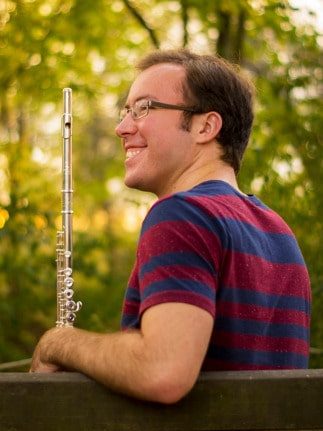 David sitting on a bench holding a flute, smiling.